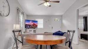 Una sala de estar moderna con una mesa redonda de madera, dos sillas, un ventilador de techo, un televisor y un reloj en la pared.