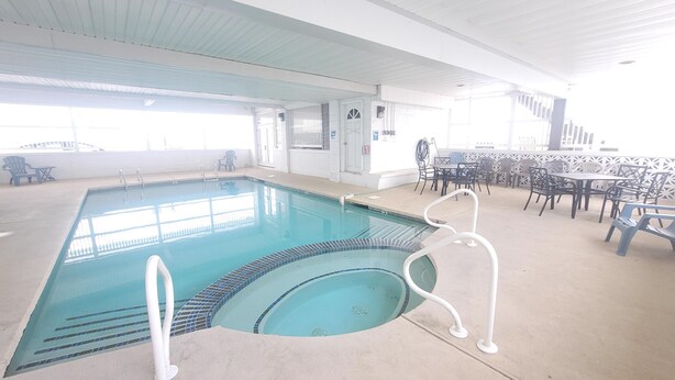 Une piscine intérieure dont les bords sont recouverts de carreaux, entourée de chaises longues et de tables.