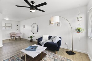 A modern living room with a dark blue sofa, a white coffee table, a floor lamp, a hanging plant, and a dining area with a table and chairs. - Contemporary Condo in Central Biltmore (Phoenix)