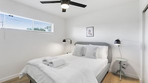 A bedroom with a bed, a ceiling fan, a bedside table, a lamp, and a window with blinds. - Contemporary Condo in Central Biltmore (Phoenix)
