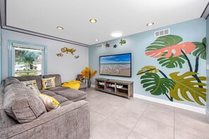 A living room with a grey sofa, a TV, a wall mural, and a window with a view of a tree. - Hibiscus16 Heated Pool/Beach/Pickleball/Tennis (Largo)