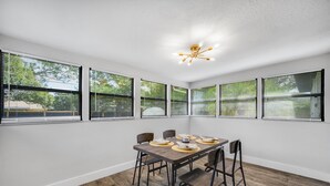 Un comedor con mesa y sillas de madera, ventanas amplias con persianas y una lámpara en el techo.