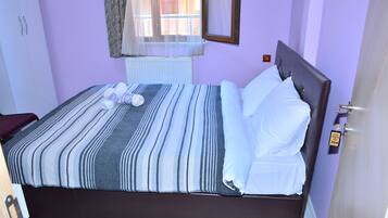 Una cama bien hecha con ropa de cama a rayas y almohadas blancas, una ventana con cortinas traslúcidas y una puerta con un cartel del número de habitación.