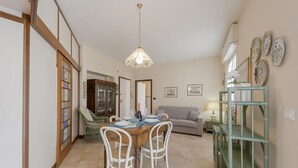 A dining area with a wooden table, white chairs, a hanging light fixture, and a green shelving unit. - GuestHost - Comfortable two-room apartment perfect for 4 guests, it is ideal for a family or small groups of friends for a relaxing holiday. We are a few minutes walk from the sea, the city center and the train station. The accommodation is equipped with (Sanremo)