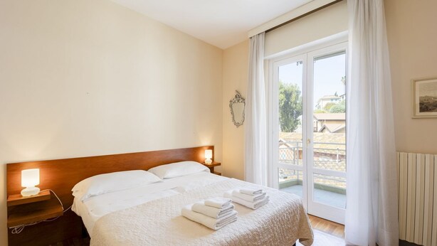 A hotel room with a bed, a nightstand, a lamp, a window with curtains, and a view of the outside. - GuestHost - Comfortable two-room apartment perfect for 4 guests, it is ideal for a family or small groups of friends for a relaxing holiday. We are a few minutes walk from the sea, the city center and the train station. The accommodation is equipped with (Sanremo)