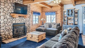 A log cabin interior with a stone fireplace, a television mounted on the wall, a ceiling fan, and a blue cabinet with shelves. - Mountain Retreat | Fast Internet | Full Kitchen (Heber City)