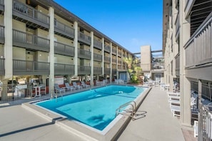 A swimming pool with lounge chairs, a covered area, and a multi-story building with balconies.