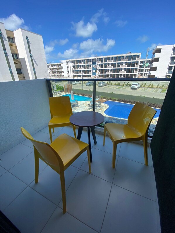A balcony with yellow chairs and a small table, overlooking a swimming pool and a parking area with buildings in the background. - Mana Beach 108D (Ipojuca)