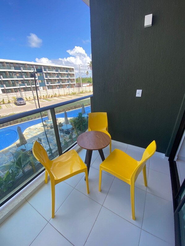 A balcony with yellow chairs and a small round table, overlooking a swimming pool and a building with multiple balconies.