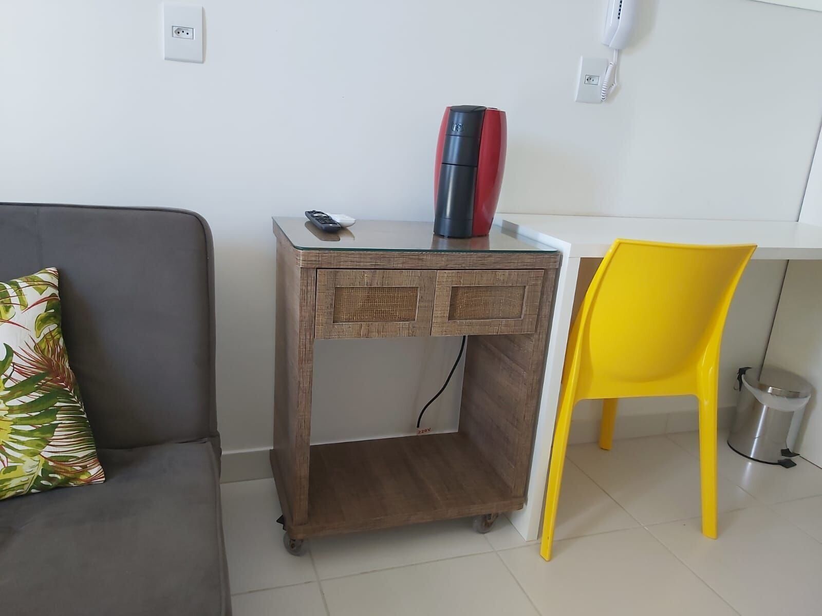 Un pequeño escritorio de madera con un calefactor rojo y negro encima, una silla amarilla y un sofá gris con un cojín estampado con hojas.