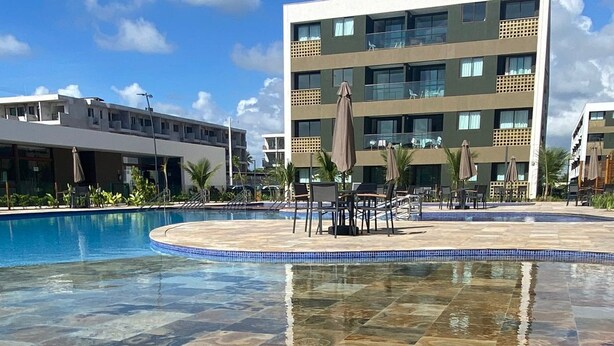 Una piscina con solárium empedrado, rodeada de sillones de baño y sombrillas, y un edificio moderno al fondo.