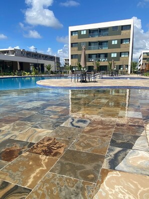 A swimming pool with a tiled deck, surrounded by lounge chairs and umbrellas, and a modern building in the background. - Mana Beach 108D (Ipojuca)