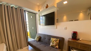 Una sala moderna con un sofá gris, un televisor de pantalla plana montado encima, una cafetera sobre una mesita de madera, y un ventanal corredero que da a un área exterior.