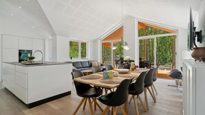 Une cuisine moderne dotée d’un îlot central, d’un coin repas avec une table en bois et des chaises noires, et de grandes fenêtres offrant une vue sur l’extérieur.