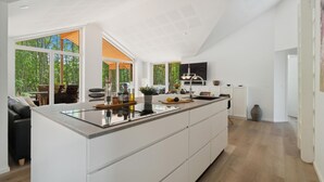Une cuisine moderne dotée d’un grand îlot central, de meubles blancs et offrant une vue sur une zone boisée grâce à de grandes fenêtres.