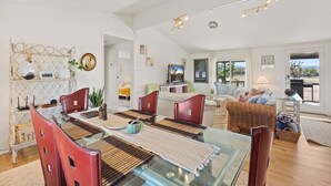 A spacious living room with a glass dining table, red chairs, and a sofa. There is a TV on a stand and a window with a view of the outdoors.