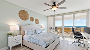 Une chambre à coucher comprenant un lit, une table de chevet, un bureau et une chaise, ainsi que de grandes fenêtres donnant sur l’océan.