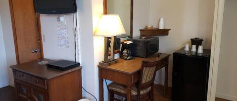 Une chambre d’hôtel de taille modeste, dotée d’un bureau en bois, d’un téléviseur fixé au mur, d’un petit réfrigérateur et d’une poubelle.