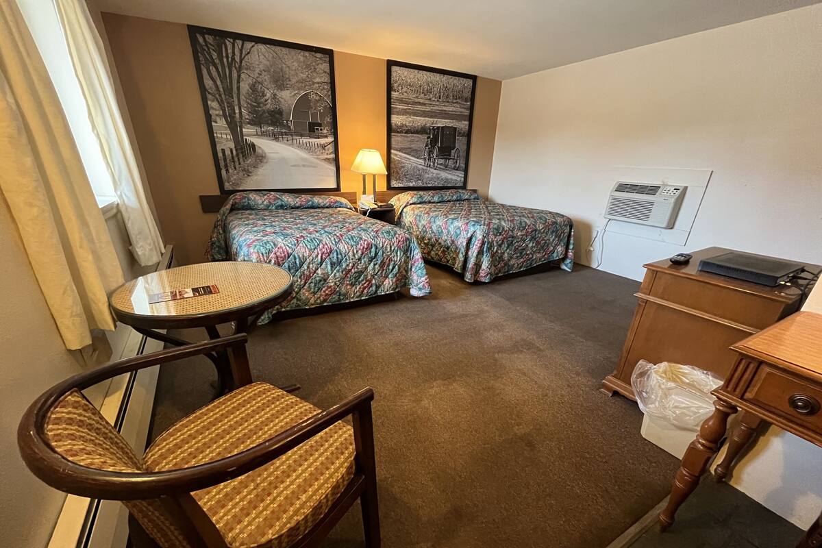 A hotel room with two beds, a small round table, a chair, a lamp, and a wooden dresser.