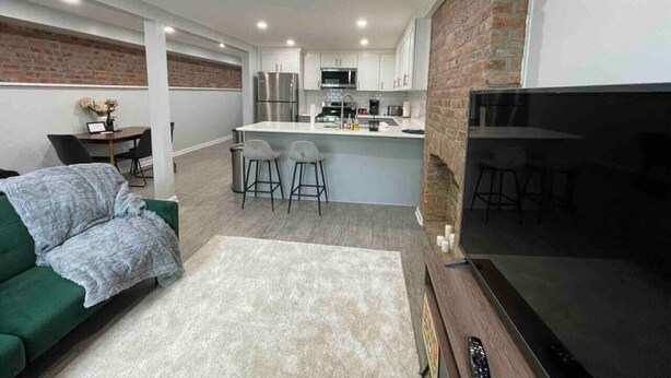 A modern living area with a green sofa, a flat-screen TV, a kitchen with white cabinets, and a brick accent wall.