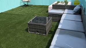 A rooftop patio with artificial grass, a wicker sofa, a glass-top table, and a hanging chair.