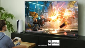 Two people playing a video game on a TV with a 'RATCHET CLANK' poster, a plant, and a 'RATING PENDING' sign.