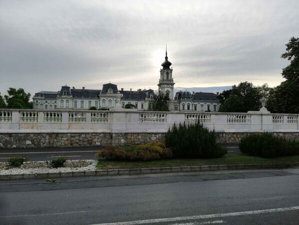 A large, historic building with a prominent tower, a stone wall with decorative balustrades, and a paved road in the foreground.