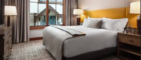 A hotel room with a large bed, a nightstand, a lamp, a window with curtains, and a view of a building outside.