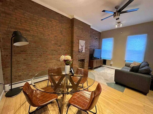 A modern living room with a brick wall, a round glass table with chairs, a sofa, a TV, and a ceiling fan.