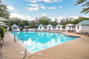 A swimming pool with lounge chairs and umbrellas, surrounded by trees and buildings with striped roofs.