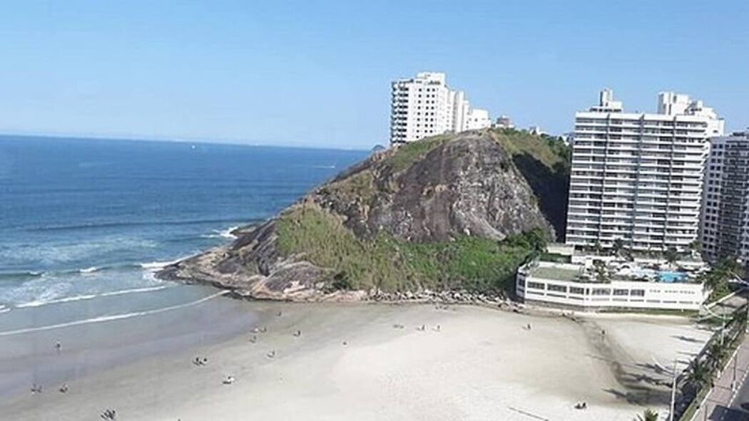 A beach with a rocky hill, a sandy shore, and several high-rise buildings in the background.