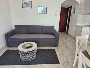 Un sofá gris, una mesa redonda de centro con base geométrica, una silla blanca y un suelo de baldosas.