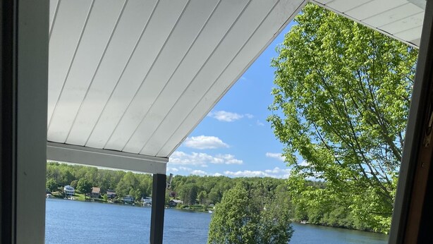 Eine überdachte Terrasse mit weißem Bodenbelag, zwei Liegestühle, ein kleiner Tisch und ein Blumentopf. Durch die offene Fläche ist ein Blick auf einen See und Grünanlagen möglich.