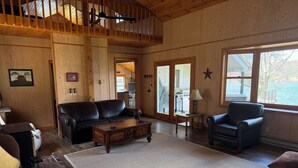 Ein geräumiges Wohnzimmer mit Holzbalkendecke, einem großen Fenster mit Blick auf ein Gewässer, einem schwarzen Ledersofa, einem Holztisch und einem schwarzen Sessel.