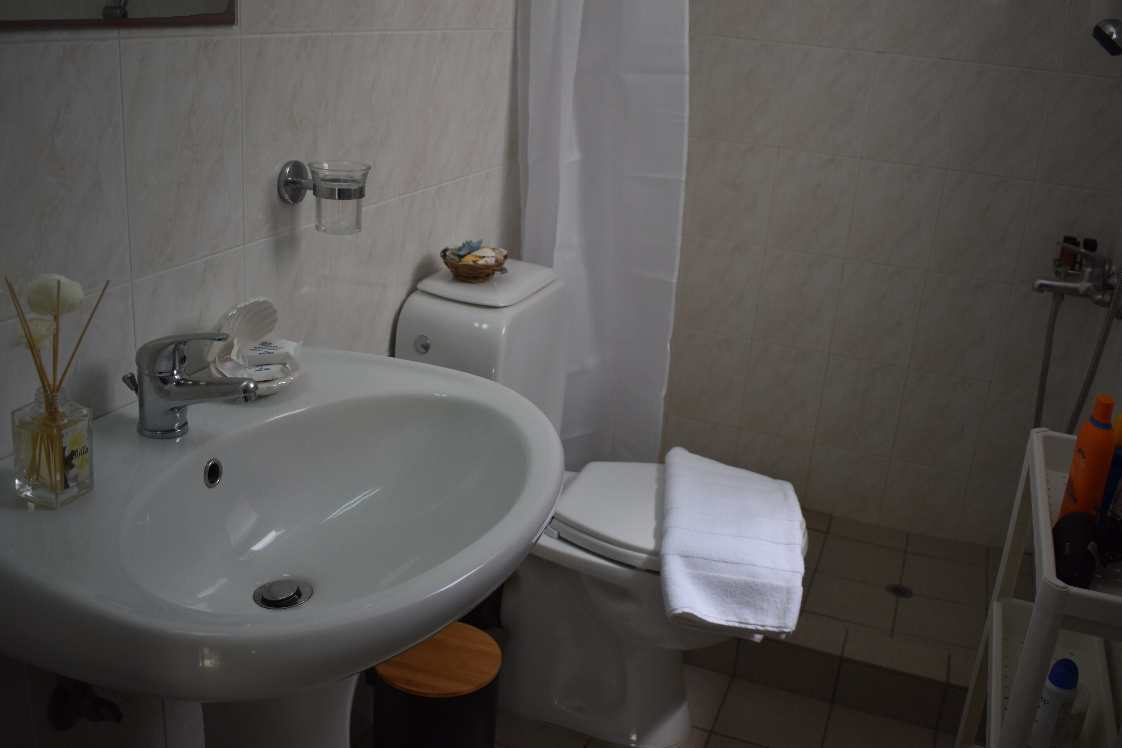 A bathroom with a white sink, toilet, and shower. There is a small basket on the toilet and a bottle of hand soap on the sink.
