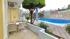 Ein Balkon mit Blick auf einen Pool, eine Essecke mit Stühlen, ein Baum und ein blauer Sonnenschirm.