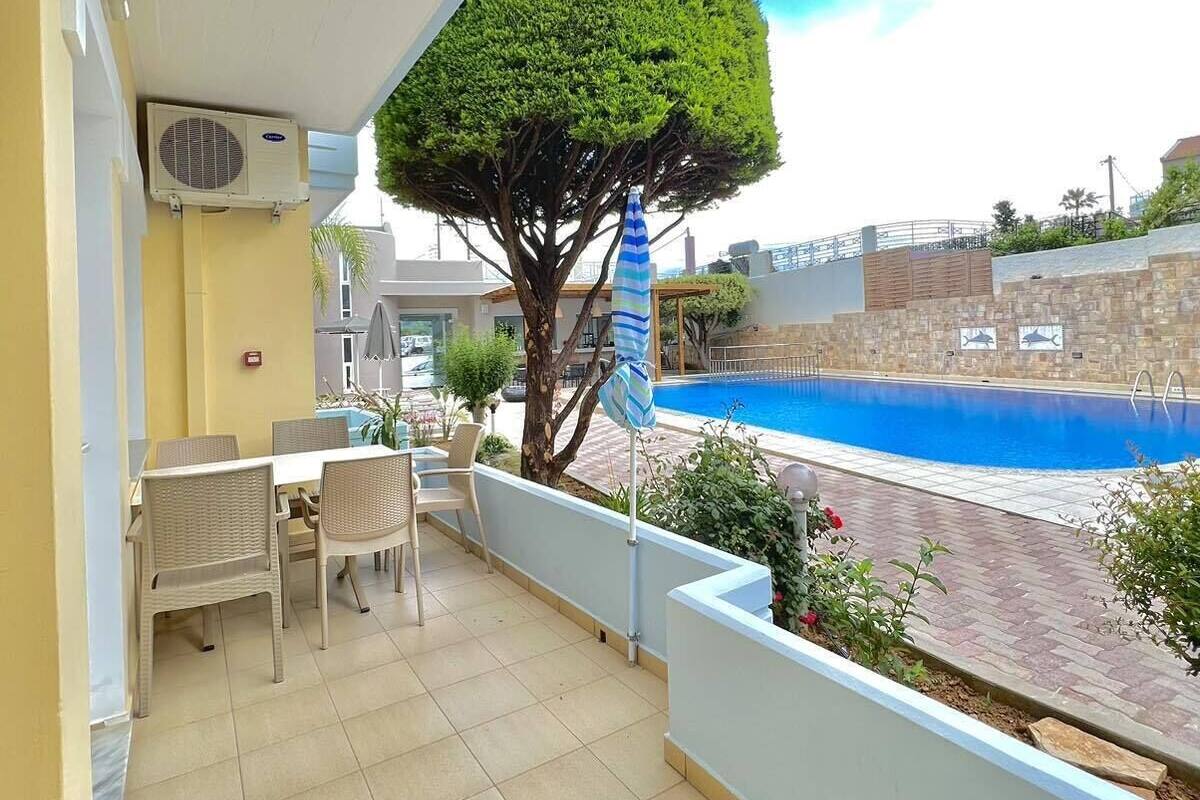 A balcony with a pool view, a dining area with chairs, a tree, and a blue umbrella.