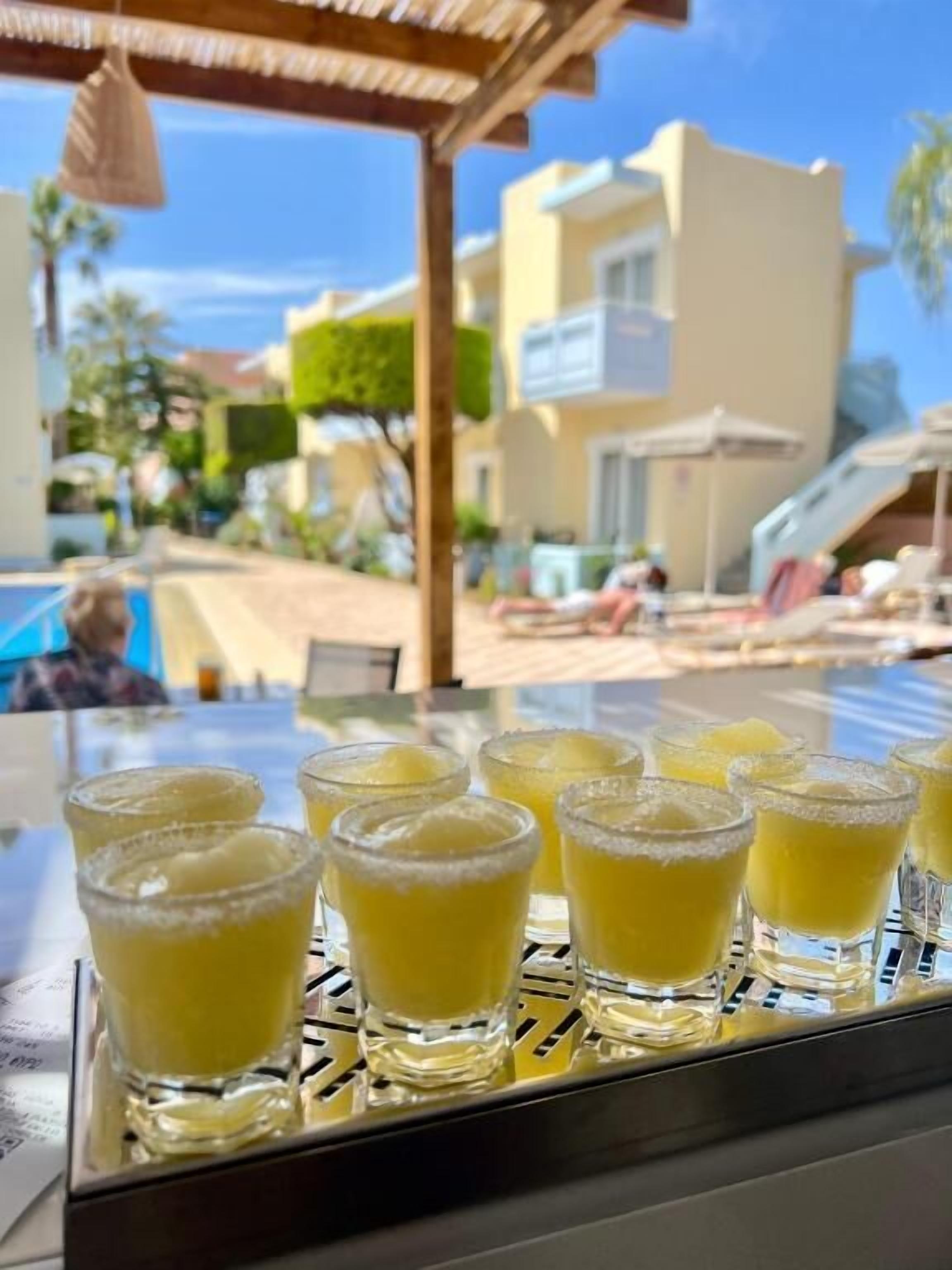 A table with several glasses of a yellow beverage, likely a cocktail, with a sugar rim. The setting appears to be an outdoor area with a pool and buildings in the background.