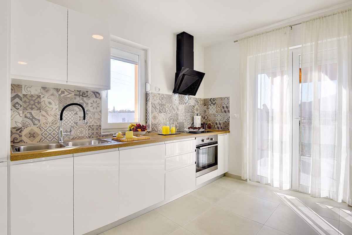 A modern kitchen with white cabinets, a stainless steel sink, a built-in oven, and patterned backsplash tiles.