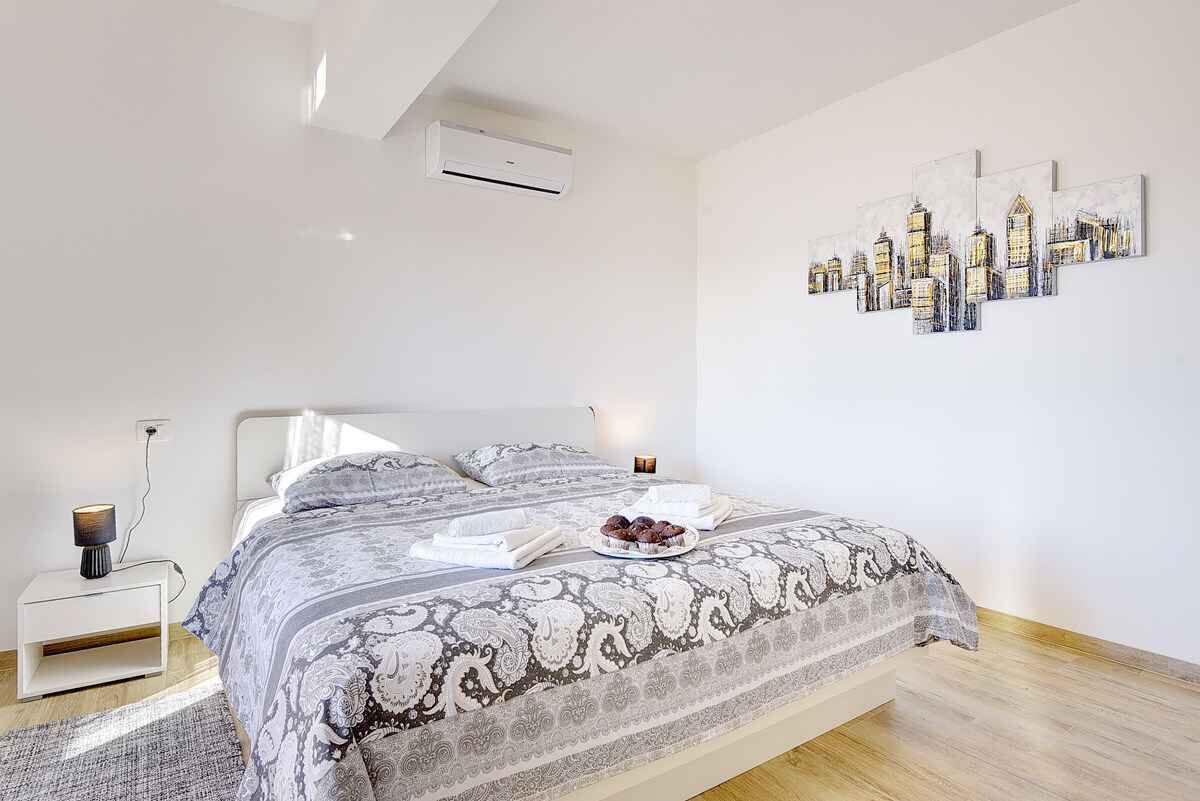 A modern bedroom with a large bed, a nightstand, a wall-mounted air conditioner, and a cityscape painting.