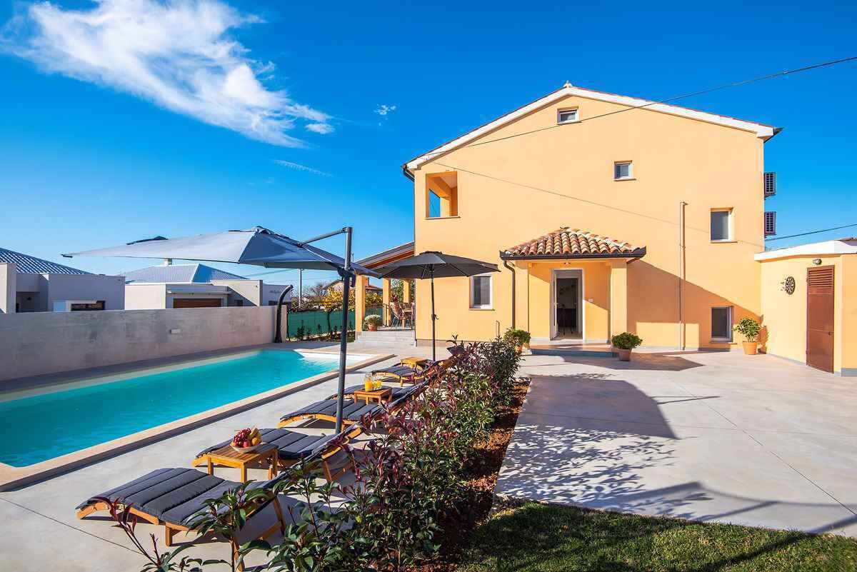 A poolside area with lounge chairs and umbrellas, a swimming pool, and a building with a tiled roof.