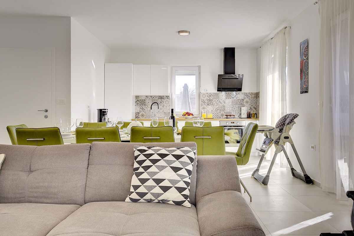 Ein modernes Wohnzimmer mit einer Couch und Sesseln, ein Essbereich mit Tisch und Stühlen sowie eine Küche im Hintergrund.