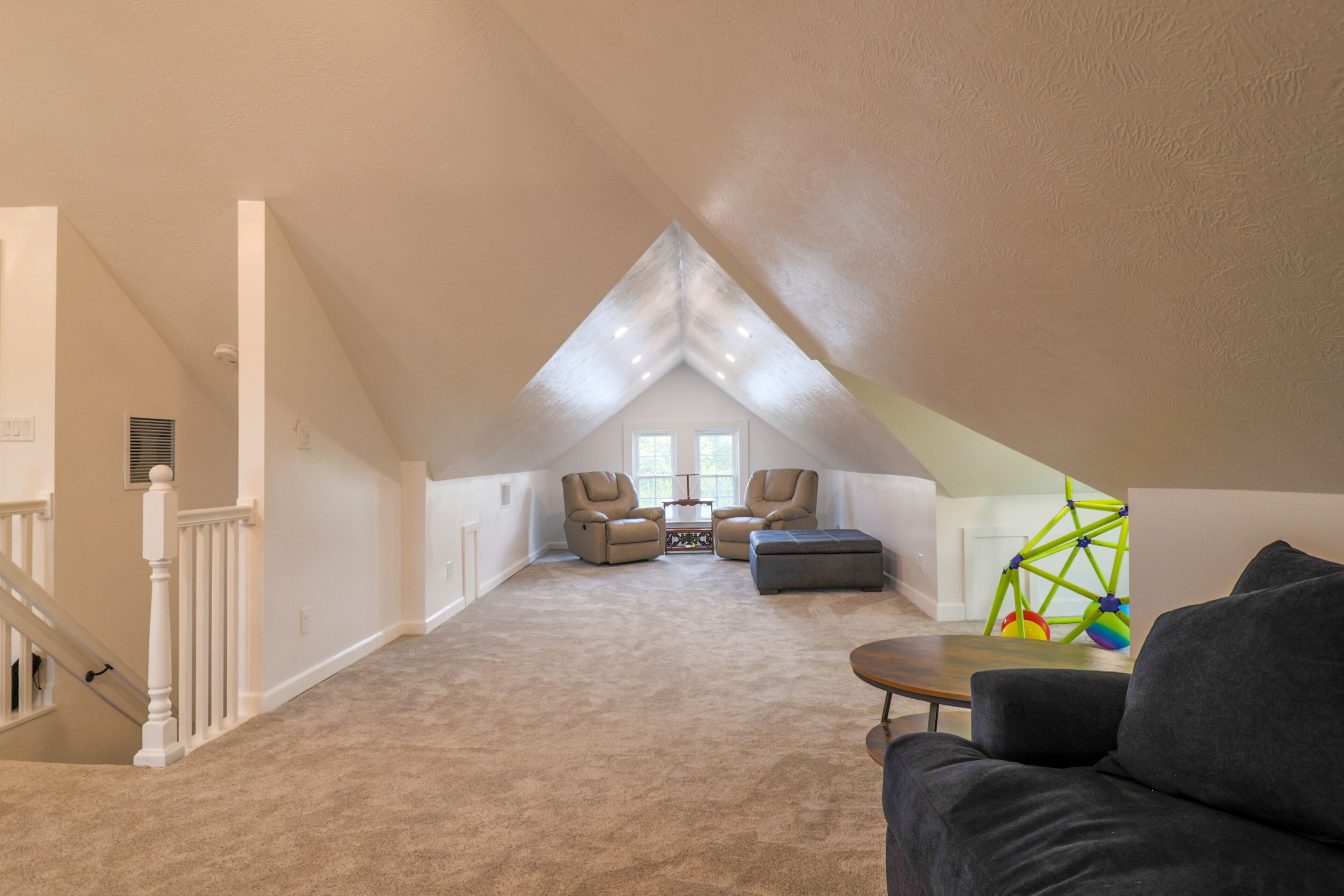A spacious attic room with a sloped ceiling, a large window, a staircase, and a seating area with a sofa and chairs.