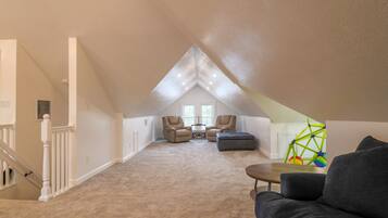 A spacious attic room with a sloped ceiling, a large window, a staircase, and a seating area with a sofa and chairs.