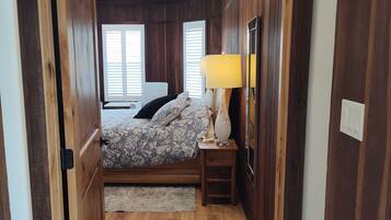 Une chambre avec un plancher en bois, un lit recouvert d’une couvre-lit à motifs floraux, une table de chevet avec une lampe et une fenêtre avec des stores.
