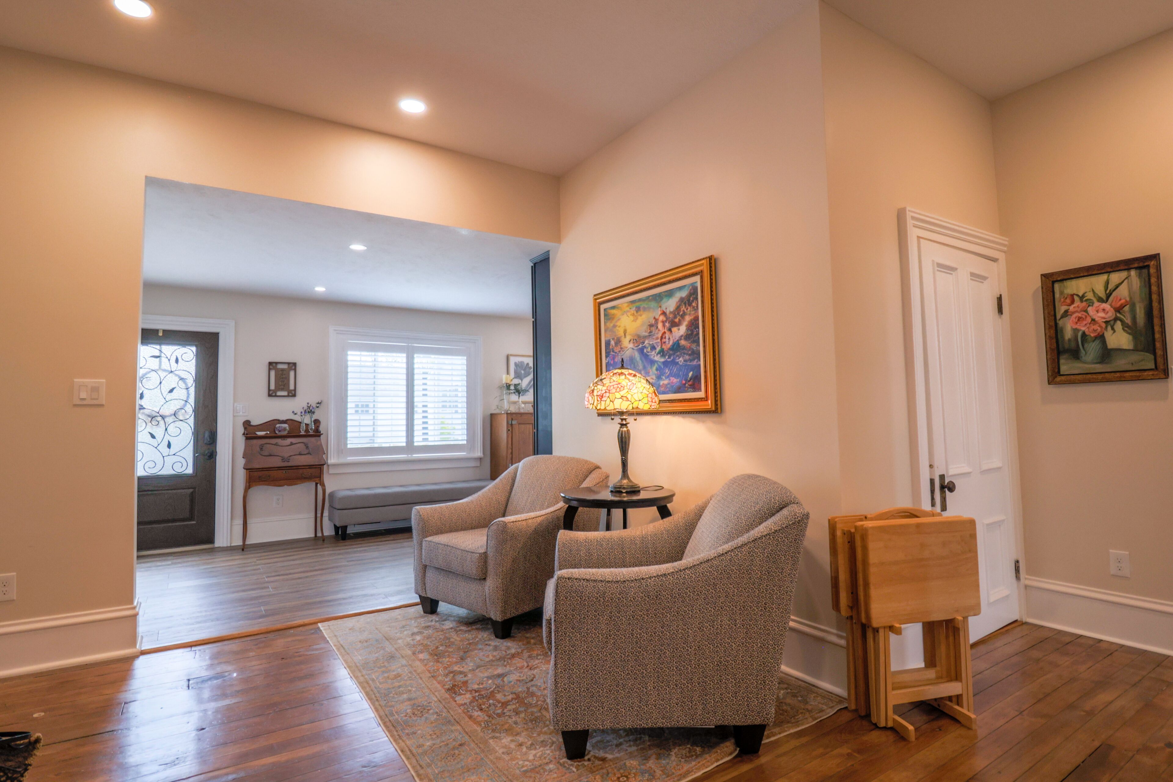 A spacious living room with hardwood flooring, two armchairs, a small table with a lamp, and a painting on the wall.