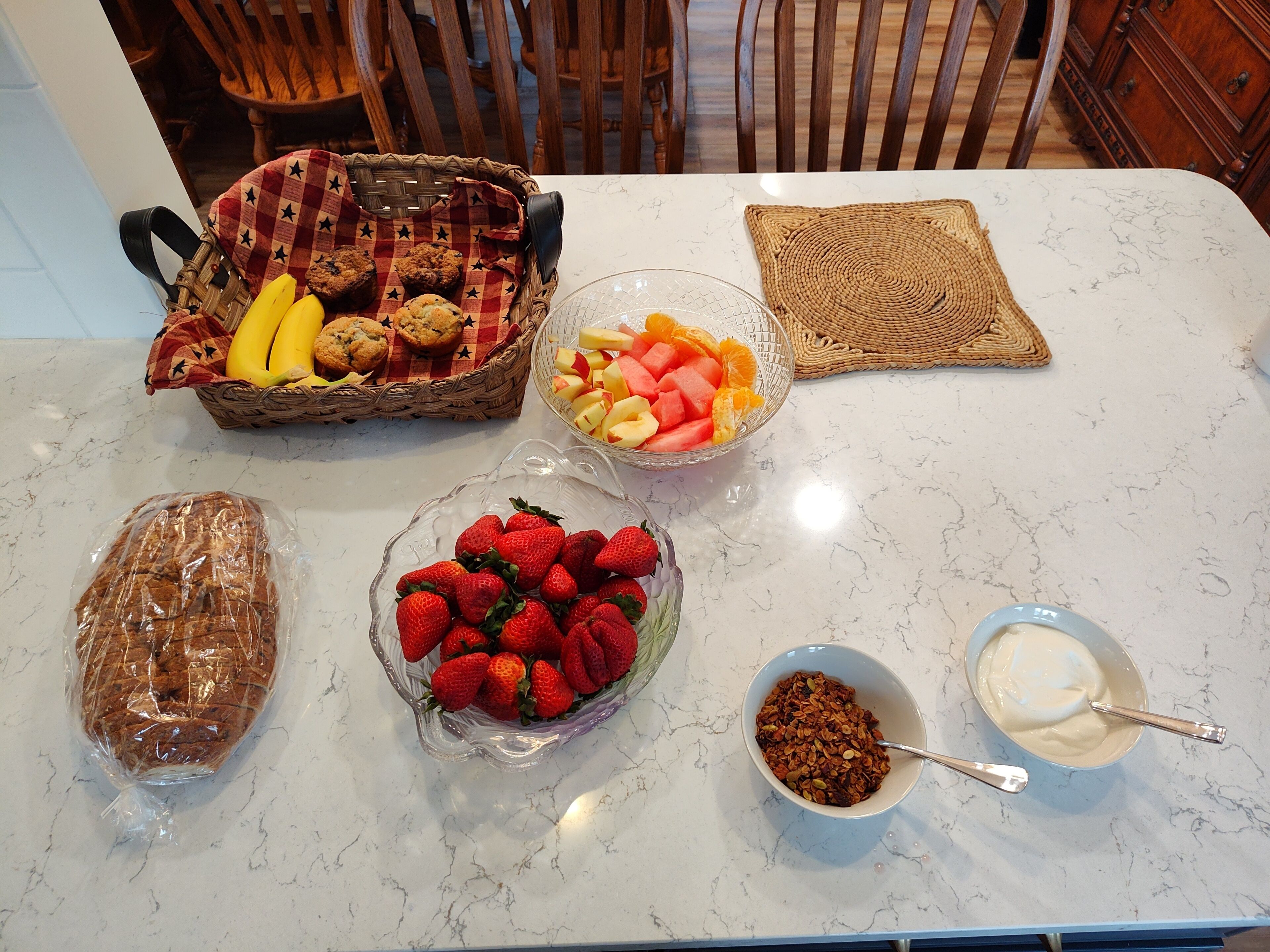 Una mesa con diversos alimentos, incluyendo un pan, un tazón de fresas, un tazón de fruta mixta, una cesta de muffins y un tazón de yogur.