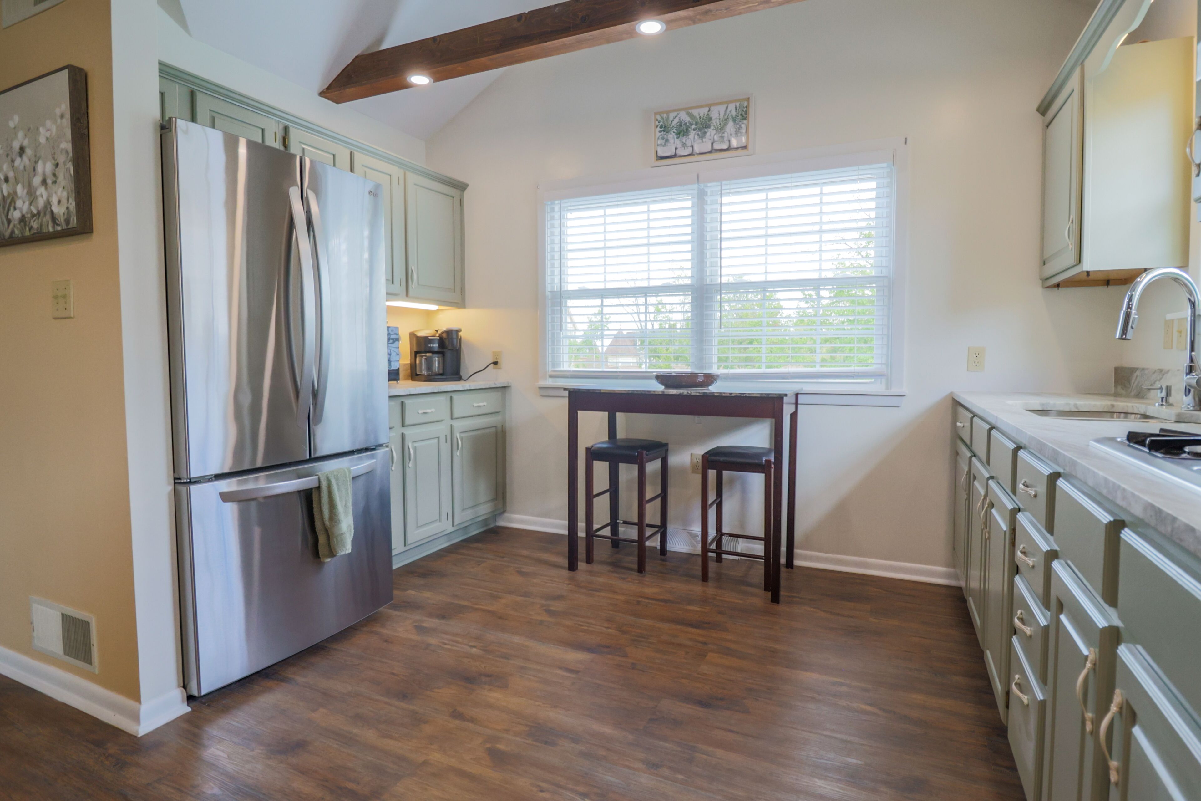 A kitchen with a stainless steel refrigerator, wooden stools, a window with blinds, and light-colored cabinets.