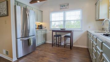 A kitchen with a stainless steel refrigerator, wooden stools, a window with blinds, and light-colored cabinets.