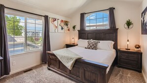 A bedroom with a large bed, a ceiling fan, a window with curtains, and artwork on the walls.